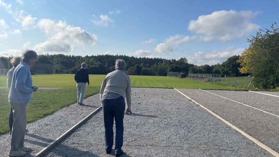 Fyra seniorer spelar boule. En kvinna ska precis kasta sitt klot. Vädret är soligt.