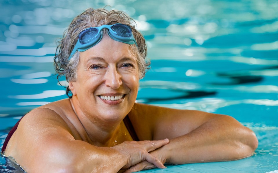 En äldre kvinna med simglasögon i pannan, som hänger på kanten av en simbassäng.